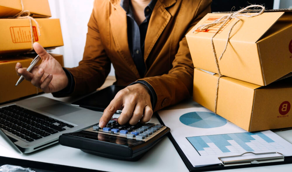 Pessoa à mesa do escritório com calculadora na mão e diversos pacotes ao lado.